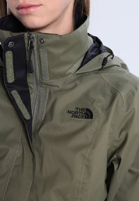 Olivgrüne wasserdichte Jacke mit hohem Kragen, schwarzen Akzenten und Reißverschluss. Mit Logo auf der Brust und strukturierten Einsätzen.
