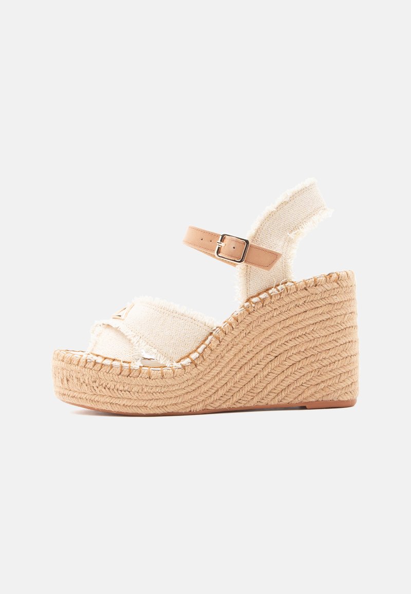 Sandale compensée beige avec plateforme tissée, brides en tissu effiloché et boucle réglable à la cheville sur fond blanc.