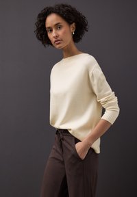 Beige gebreide trui met een ronde hals en lange mouwen, gecombineerd met donkerbruine broek met zakken en een aantrekkoord in de taille.