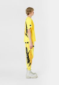 Camiseta amarilla con un gráfico de alas negras en la parte posterior, combinada con pantalones de chándal amarillos a juego con patrones y franjas laterales negras. Botas grises.