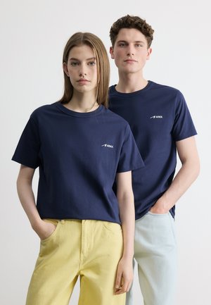 Jauna moteris ir vyras stovėdami dėvi atitinkančius tamsiai mėlynus ARKK marškinėlius; moteris vilki geltonas kelnes, vyras – šviesiai mėlynas kelnes, abu rankas kišenėse.