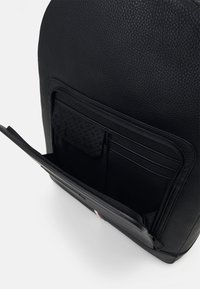 Sac à dos en cuir noir avec une finition texturée, poche zippée et plusieurs emplacements pour cartes à l'intérieur. Présente un petit accent coloré à l'avant.