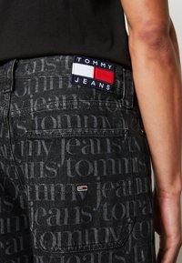 Crne traperice od denima s suptilnim tonalnim uzorkom "tommy" teksta. Imaju brendiranu oznaku na pojasu i mali logo na stražnjem džepu.