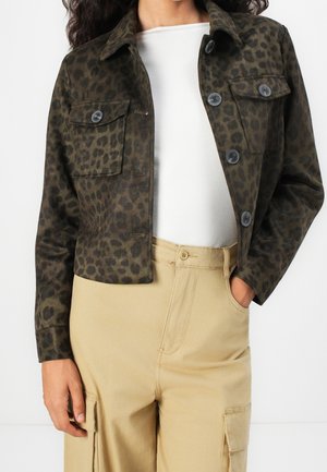 Kvinde iført mørkegrøn leopardmønstret cropped jakke over hvid top og beige højtaljede cargobukser med lommer og knapper.