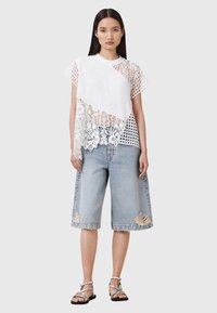 White lace and mesh top with an asymmetrical design, paired with light blue distressed denim culottes. Model wearing silver sandals.