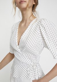 Donna che indossa una blusa avvolgente bianca con pois neri, maniche a sbuffo e una cinta in vita, abbinata a pantaloni bianchi.