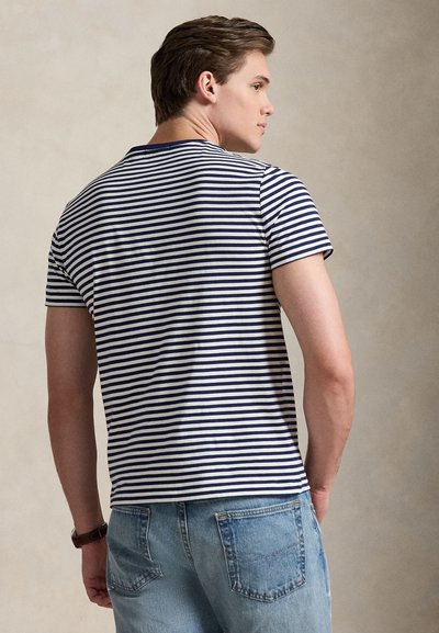 Νεαρός άνδρας που φοράει κοντομάνικο μπλουζάκι με ναυτικό μπλε και λευκές ρίγες και ανοιχτόχρωμα τζιν, γυρισμένος ελαφρώς στο πλάι μπροστά σε ουδέτερο φόντο.