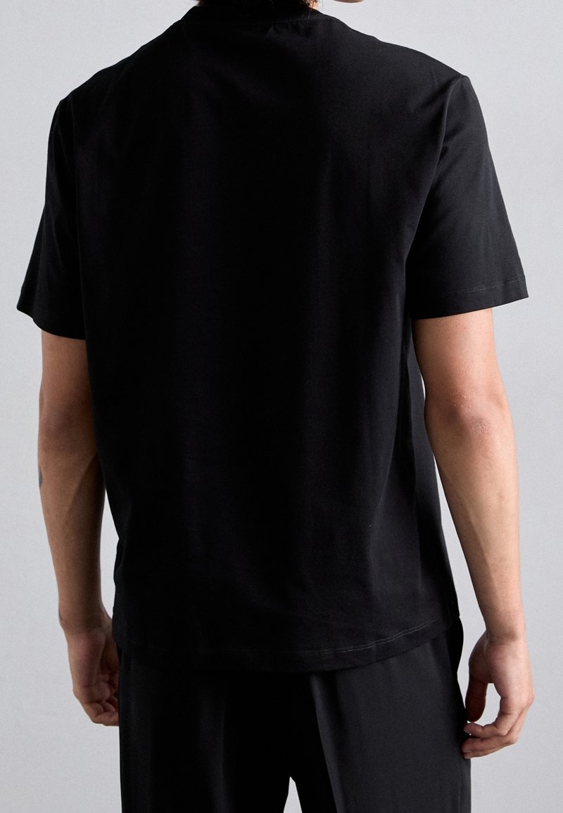 Personne vue de dos portant un t-shirt noir uni à manches courtes et un pantalon noir sur un fond gris neutre.