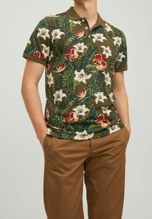 Homme portant un polo manches courtes vert olive avec imprimé floral tropical et pantalon marron, mains dans les poches, sur fond uni.