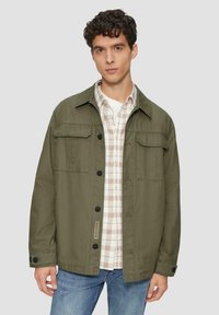 Olijfgroene jas met knoopsluiting, twee borstzakken en overhemdkraag, gelaagd over een geruit shirt, gedragen met blauwe spijkerbroek.