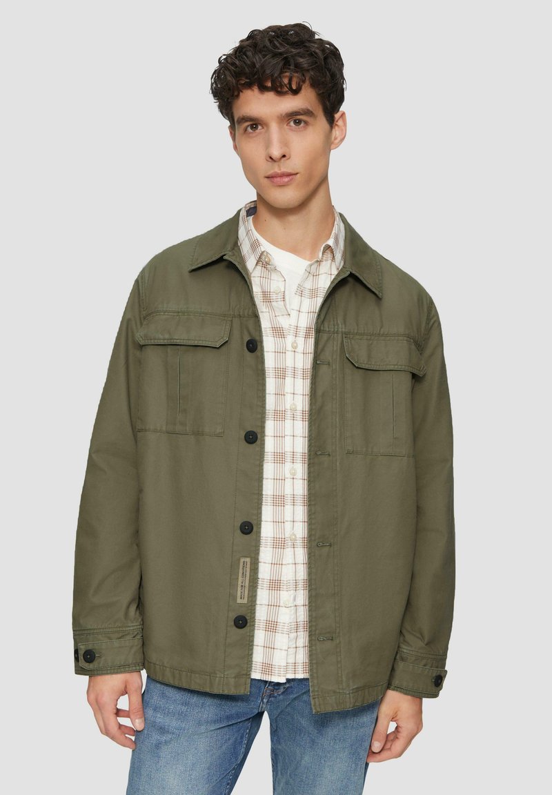 Olijfgroene jas met knoopsluiting, twee borstzakken en overhemdkraag, gelaagd over een geruit shirt, gedragen met blauwe spijkerbroek.