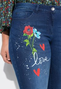 Jeans de mezclilla azul oscuro con gráficos florales vibrantes en rojo y azul, y la palabra "amor" en escritura blanca con acentos salpicados.