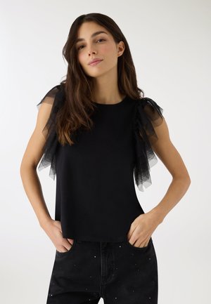 Kobieta z długimi brązowymi włosami, ubrana w czarny top z przezroczystymi, falbanianymi rękawami oraz czarne spodnie z drobnymi białymi kropkami, stoi z rękami w kieszeniach na białym tle.