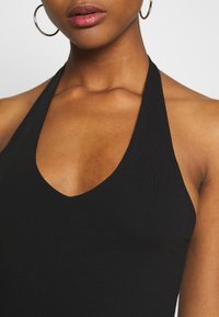 Černý halter top s hlubokým výstřihem ve tvaru V, vyrobený z hladké látky. Disponuje přiléhavým střihem a stylem s odhalenými rameny. Viditelné kruhové náušnice.