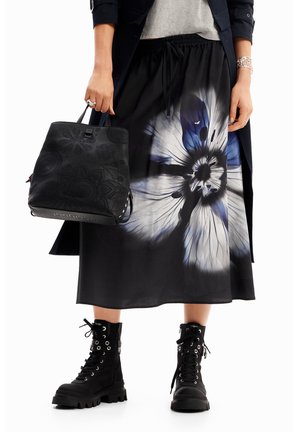 Femme portant une jupe midi noire à fleurs, des bottes noires à lacets, un sac à main noir à motifs, et des bracelets argentés.