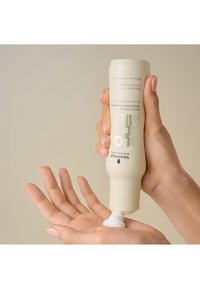 Swanwood Bond Repair Conditioner in een zachte witte tube, die romige product uitperst op een lichtgetinte palm, matte afwerking, minimalistisch ontwerp.