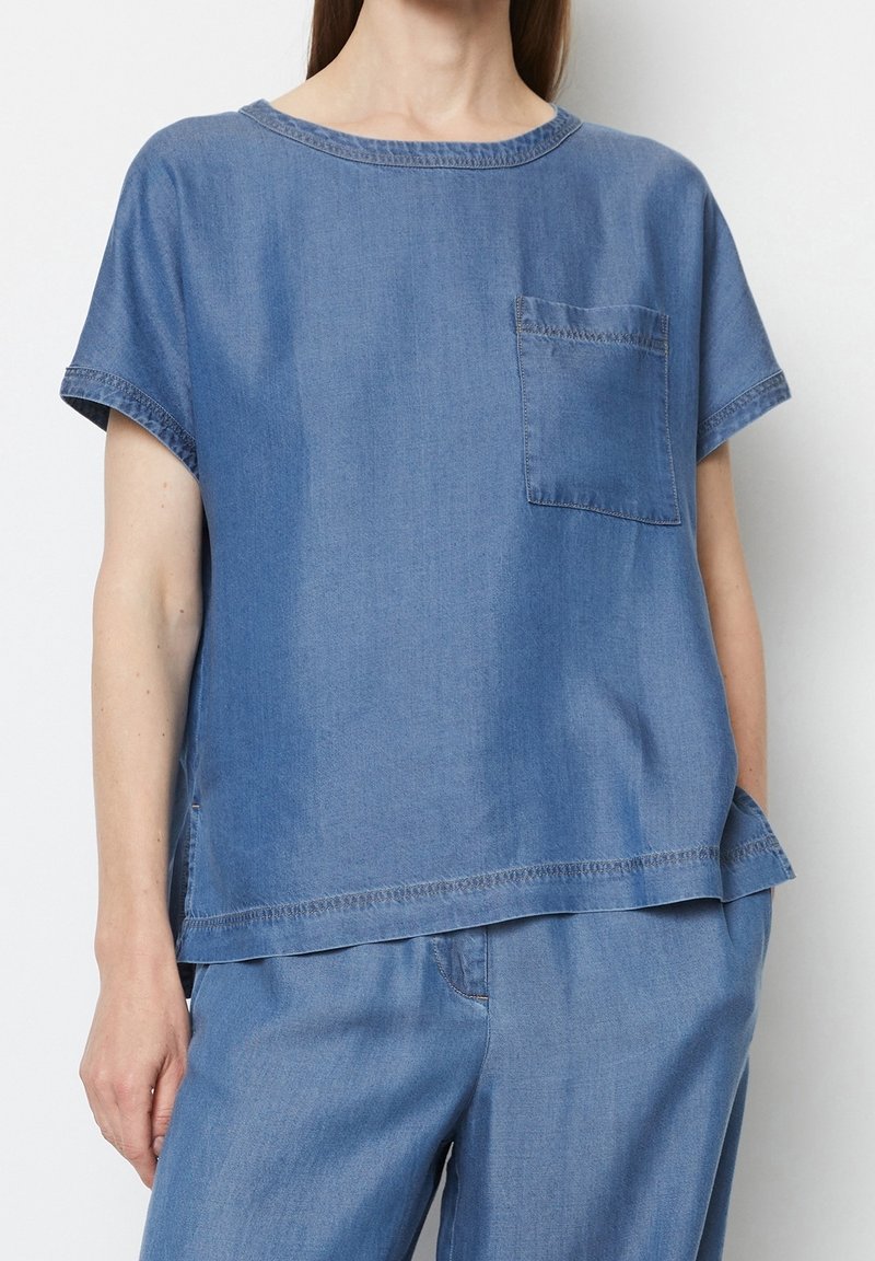 Top in denim di colore azzurro chiaro con maniche corte, scollatura rotonda e taschino frontale. Presenta una vestibilità morbida e dettagli di cucitura in evidenza.