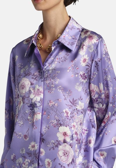 Woman wearing a lavender satin blouse with white and purple floral pattern and a gold twisted chain necklace, against a white background.