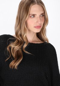 Pulover negru din tricot cu un guler larg și lejer și o țesătură texturată. Păr lung și ondulat care cade pe umeri. Fundal neutru.