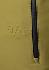 Borsa in nylon verde con una texture liscia, zipper nera, logo "a/u" in rilievo e linee pulite. Design compatto ideale per l'uso quotidiano.