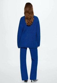 Blauwe gebreide trui met een losse pasvorm en verlaagde schouders, gecombineerd met wijde broekspijpen, beide met een gestructureerde afwerking. Zwarte hakken zichtbaar.