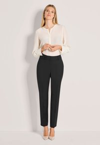 Weiße Bluse mit langen Ärmeln und einem Ausschnitt, kombiniert mit maßgeschneiderten schwarzen Hosen. Das Modell trägt nudefarbene High Heels.