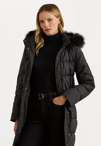 Čierny puffový kabát s umelou kožušinou na kapucni, so zipsom a patentkami, prešívanou textúrou a bočnými vreckami, v kombinácii s čiernym rolákom a džínami.