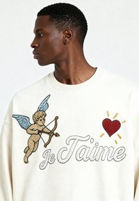 Jeune homme en sweat blanc avec un cupidon brodé tenant un arc, un cœur rouge, et le texte "Je T'aime" sur la poitrine, tourné vers la gauche sur un fond uni.