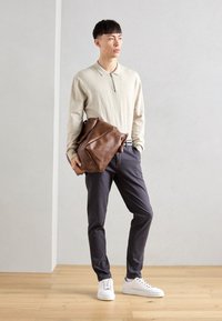 Beige langärmige polo met een rits, donkergrijze broek en witte sneakers. Bruine leren tas met een gestructureerd oppervlak in hand gehouden.