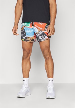 Mann trägt mehrfarbige Nike-Laufshorts, weiße Nike-Socken und weiße Sportschuhe und steht vor einem einfarbigen Hintergrund.