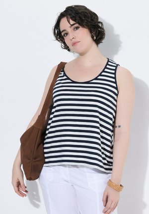 Junge Frau mit kurzen lockigen Haaren, die ein schwarz-weiß gestreiftes Tanktop, weiße Hosen, eine braune Umhängetasche und ein Armband trägt.