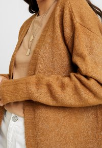 Bronsfärgad stickad kofta med öppen front, ribbade ärmslut och texturerat tyg. Lager på en beige topp med knappdetalj.