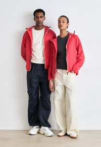 Chaquetas rojas con cremallera, capas base blancas y azul marino, pantalones anchos y ligeros; calzado deportivo con detalles texturizados y contrastes de color.