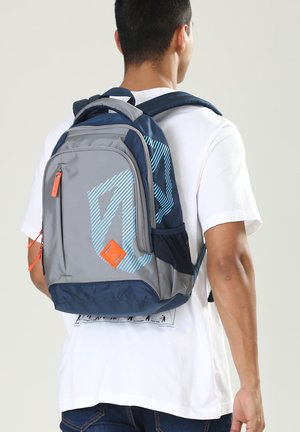 Giovane uomo che indossa uno zaino grigio e blu navy con strisce diagonali blu e tiretti arancioni, visto di spalle.