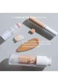 Trois produits de maquillage sur un fond gris : un enlumineur en bâton transparent, un correcteur liquide marron et un fond de teint beige avec un emballage blanc.