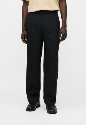 Bărbat purtând pantaloni negri cu picior drept, pantofi negri din piele și o cămașă bej cu mâneci scurte, stând în fața unui fundal gri simplu.