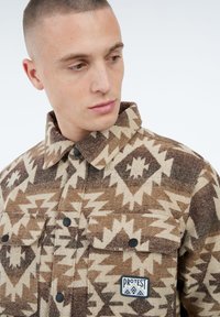 Jonge man met kort haar, gekleed in een bruine en beige gestreepte overhemdjasje met zakflappen en een "Protest" patch.