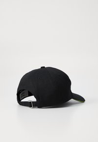 Svart bomull baseball cap med buet skygge. Har en justerbar stropp bak og en grønn detalj under skyggen.
