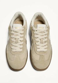 Beige Wildledersneaker mit weißen Akzenten, runder Zehenpartie und flachen Schnürsenkeln. Verfügt über eine gepolsterte Knöchelpartie und eine strukturierte Gummi-Sohle.