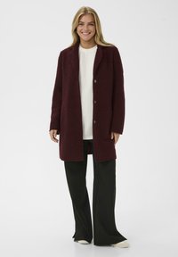 Burgundska volnena plašča s ovratnikom, odprto spredaj in dvema gumboma. Nosi se nad belim vrhom in v kombinaciji s črnimi širokimi hlačami. Gladka tekstura.