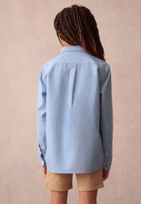 Vaaleansininen pitkähihainen nappipaita, jossa on takana laskos. Yhdistetty khakinvärisiin shortseihin; pehmeä kangas ja klassinen kaulussuunnittelu.