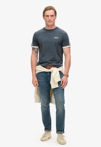 Camiseta de algodón azul marino con mangas acanaladas blancas, sudadera beige clara atada a la cintura, vaqueros azules y zapatillas color crema.