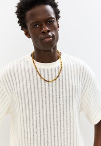 Maglione bianco a costine con maniche corte, completato da una collana di perline multicolori con accenti gialli e marroni.