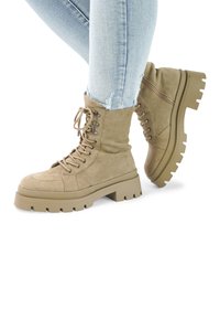 Tan suede lace-up ankle boots with a chunky, ridged sole. Features metal eyelets and a padded collar. Worn with light blue ripped jeans.