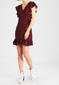 Femme portant une robe portefeuille à pois bordeaux avec des volants et des bottines blanches à talons, se tenant devant un fond uni.