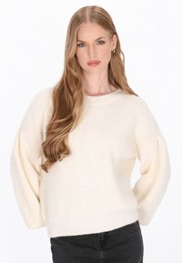 Pulover tricotat de culoare crem, cu o textură ribbed, mâneci lungi tip balloon și guler rotund. Purtați-l cu blugi negri.