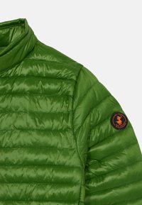 Veste rembourrée verte avec un col montant, texture matelassée, et un patch logo circulaire orange sur l'épaule. Le matériau est brillant et lisse.