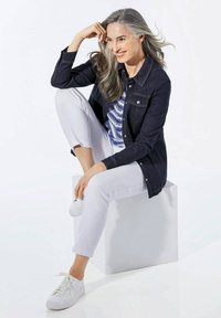 Dunkelblaue Jeansjacke über einem blau gemusterten Oberteil, weiße Knöchelhosen und weiße Sneakers. Glatte Texturen und lockerer Schnitt. Sitzpose.