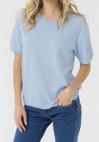 Hellblaues Strickoberteil mit Rundhalsausschnitt und kurzen Ärmeln, versehen mit strukturierten Punktmustern und lässiger Passform. Kombiniert mit blauen Jeans.