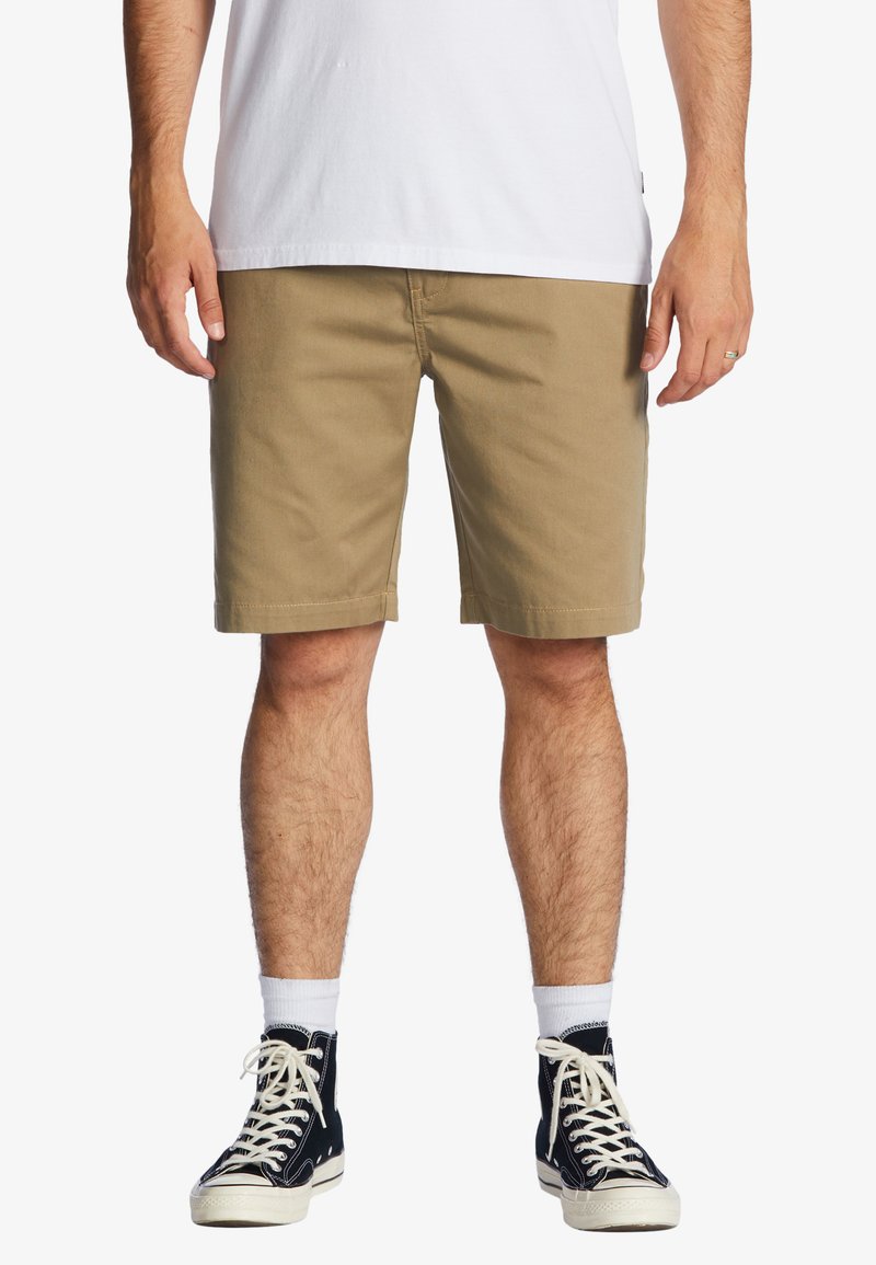 Pantaloni corti in cotone khaki con taglio dritto, fino al ginocchio, dotati di tasche laterali e chiusura a bottone; abbinati a sneakers alte blu navy.